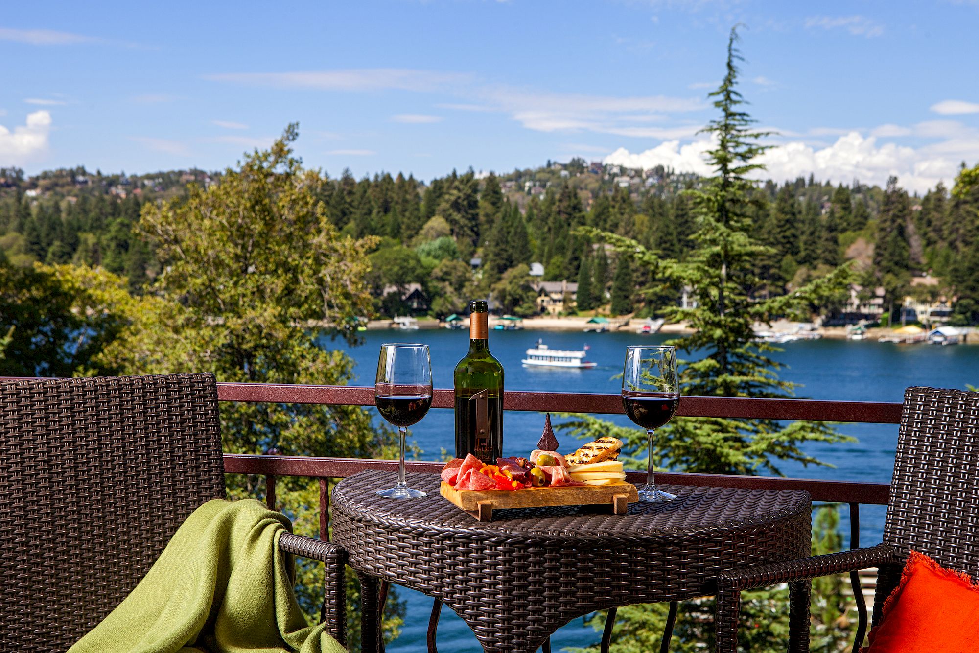 An outdoor table for two overlooks a lake, with wine bottles and glasses beside a cheese and charcuterie board.