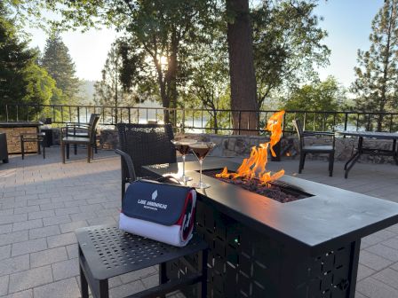 A cozy patio bonfire glows by lounge chairs, with martini glasses and a sunset sky through the trees.