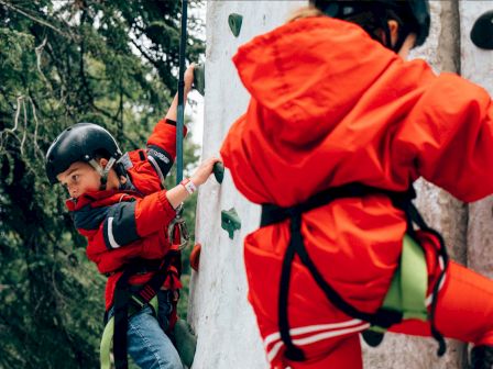 A child in a red climbing harness scales an outdoor rock wall, reaching for holds while wearing a helmet.