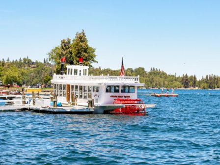 A white sightseeing boat with red accents floats on a lake near a dock, with people on the water.
