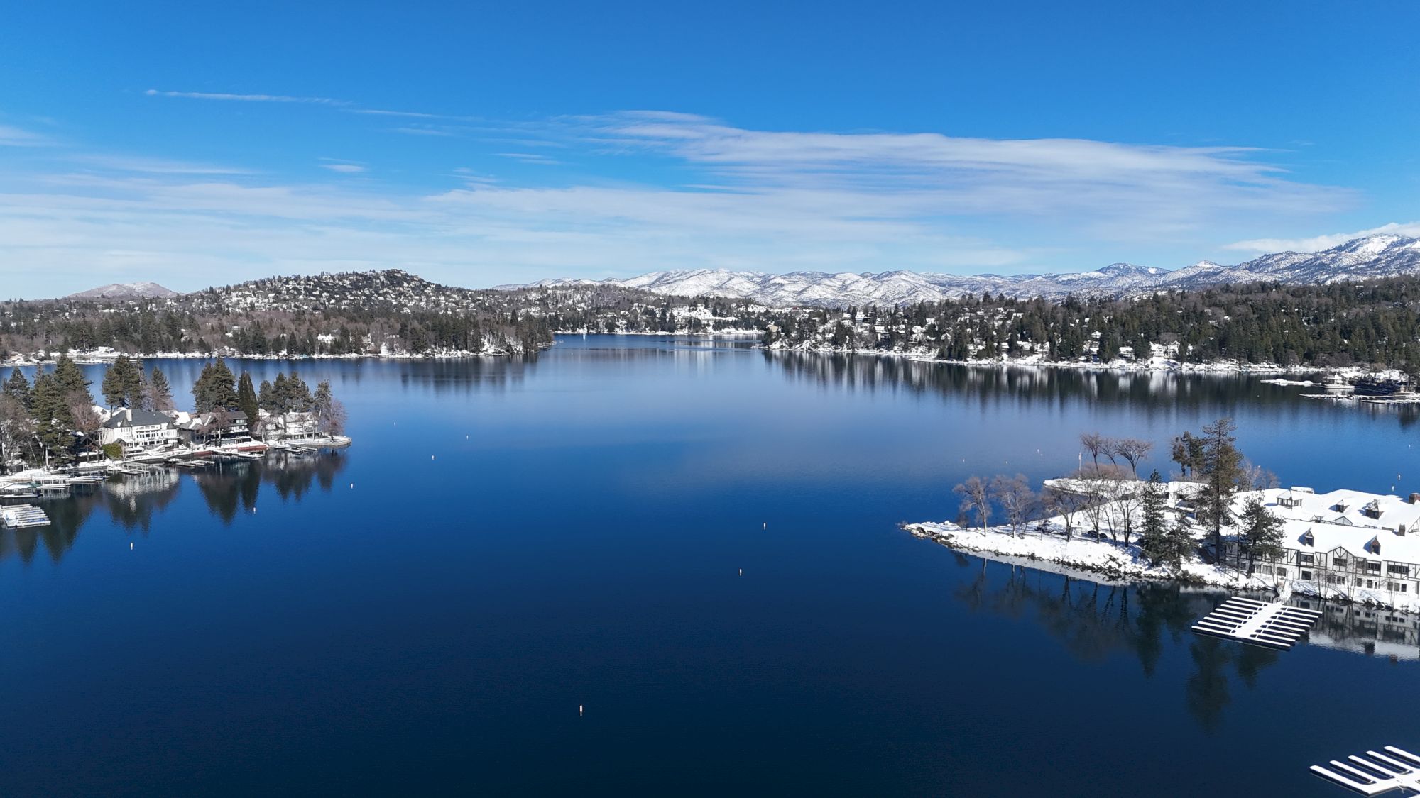 Snowy mountains and pine trees frame a calm winter lake with a riverside town and reflected rooftops, under a bright blue sky.