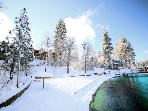Snowy riverside resort with frosted trees, covered buildings, and a bright winter sky beside green water.