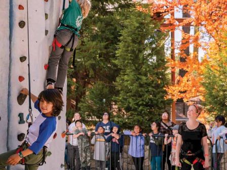 A climber scales an outdoor rock wall while spectators watch on a sunny day with orange fall trees nearby.