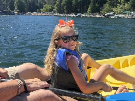 A family paddles a yellow kayak on a sunny lake, with a child in a blue life vest and orange bow watching the tree-lined shore.