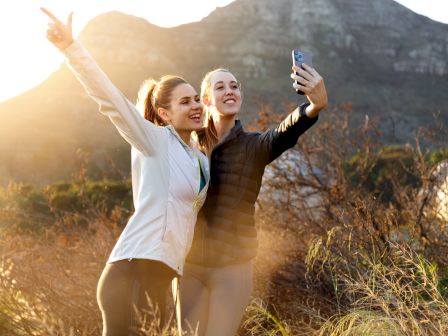 Two women in jackets take a selfie with a phone outdoors at golden sunset, smiling in front of mountains and brush.