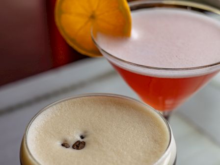 Two cocktails with frothy tops&mdash;one orange with a lemon slice&mdash;sit side by side in glasses, ready to sip.