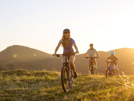 A group of cyclists rides along a grassy trail at sunset, smiling and wearing helmets.