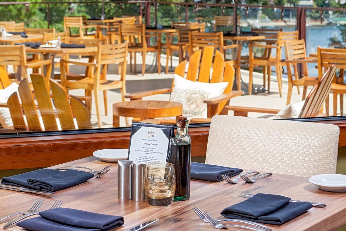 An outdoor restaurant dining area with neatly set wooden chairs and tables, plus place settings and a bottle on the table.