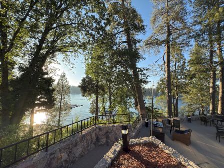 Sunlit lakeside patio with benches and caf&eacute; lights, surrounded by tall trees and a calm water view.