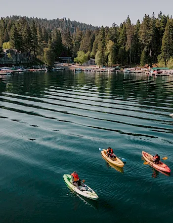 Kayakers paddle past docked boats in a calm lake, with forested mountains and lakeside buildings lining the shore.