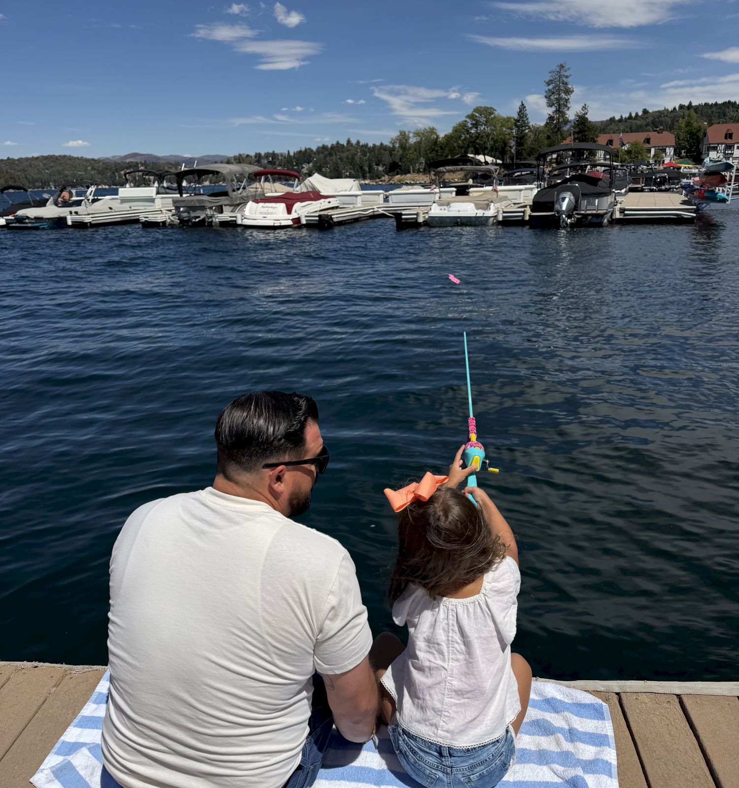 A man and a child sit by a dock on a lake, holding a bright toy water gun while boats fill the harbor behind them.