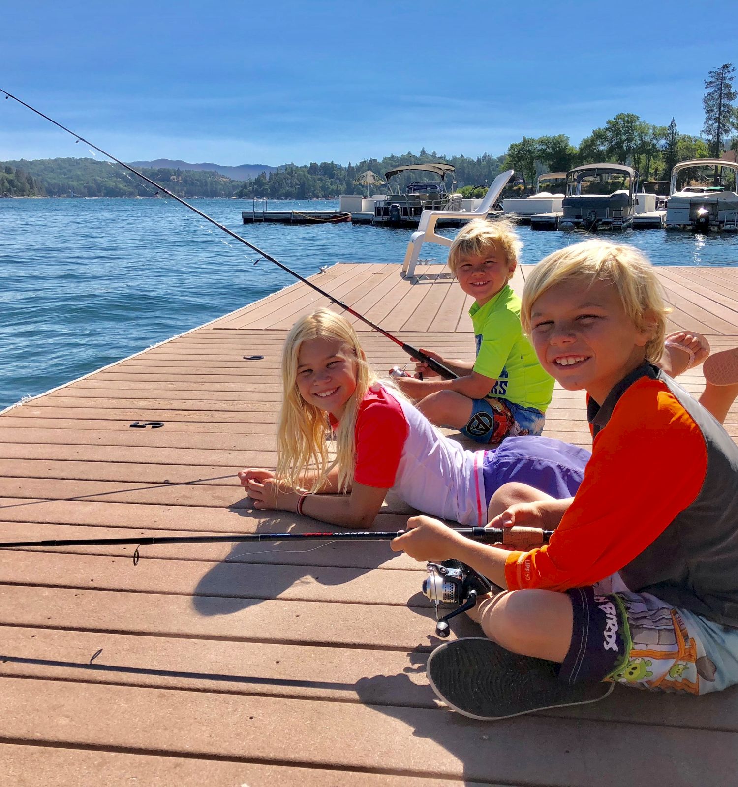 Three kids smile on a lake dock with fishing rods near moored boats under a bright blue sky.
