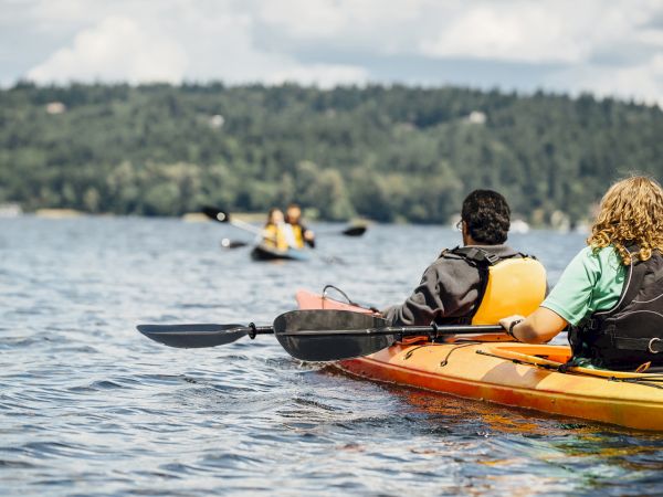 Two people in kayaks with life jackets on a lake, paddling toward a distant group of kayakers on calm water. End.