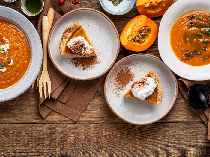 Plates of pumpkin soup and pumpkin pie slices on a rustic table, topped with cream, spices, and pumpkin seeds.