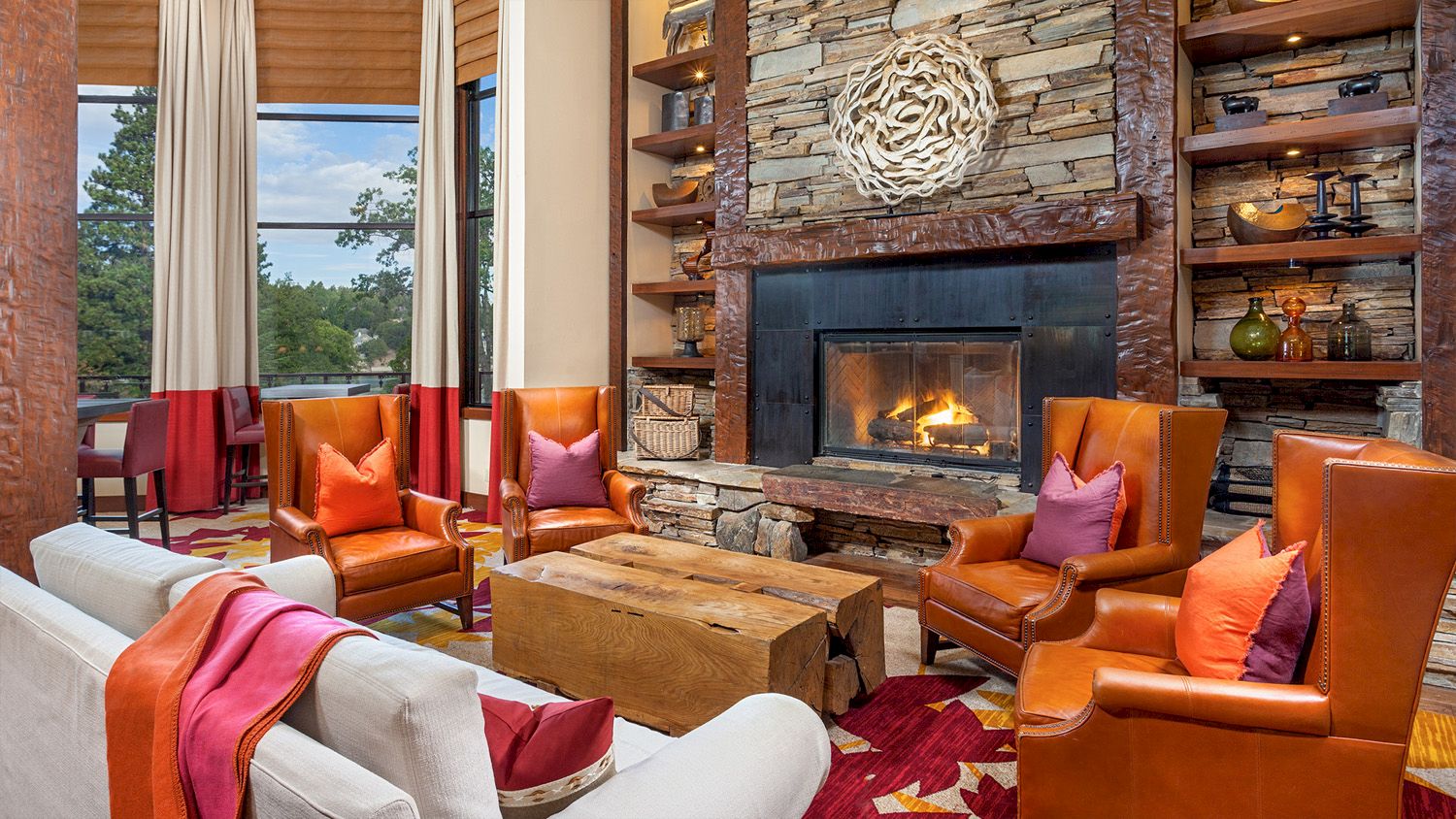 A cozy living room with a stone fireplace, warm leather armchairs, colorful pillows, and rustic wooden decor by large windows.