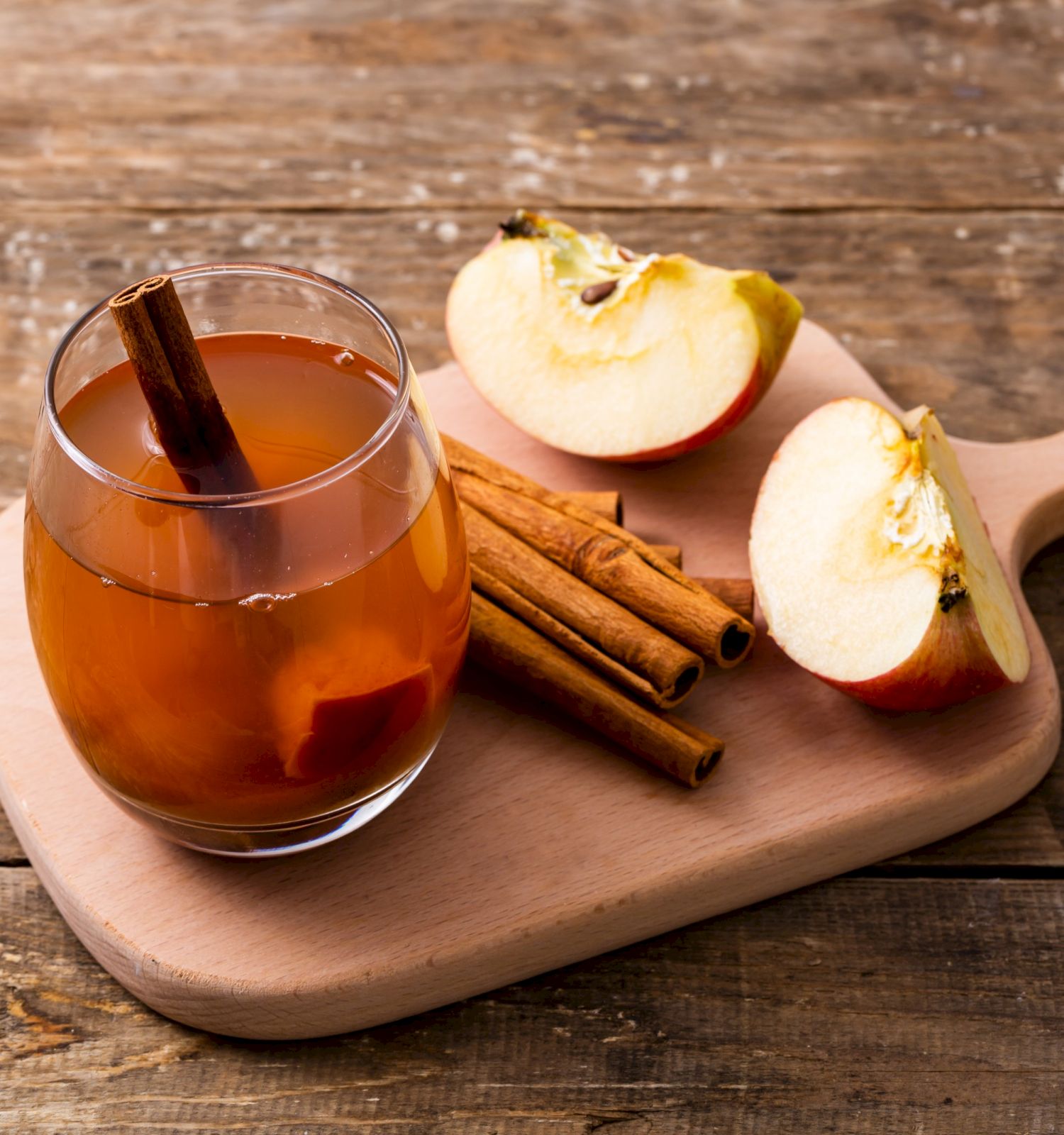 A glass of spiced apple cider with cinnamon sticks, alongside sliced apples on a wooden board.