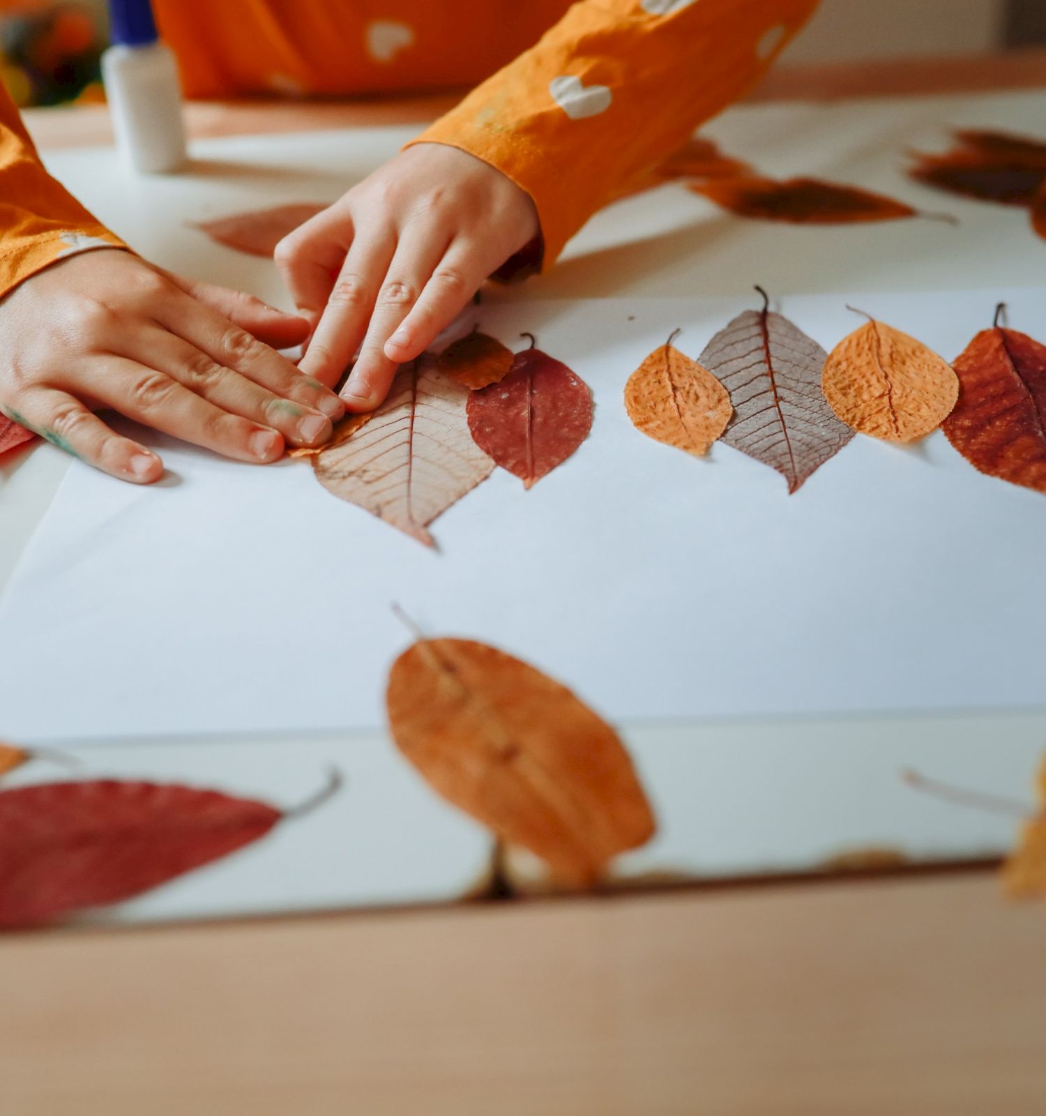A child in an orange sleeve arranges scattered autumn leaves on a table, creating a leaf craft collage.