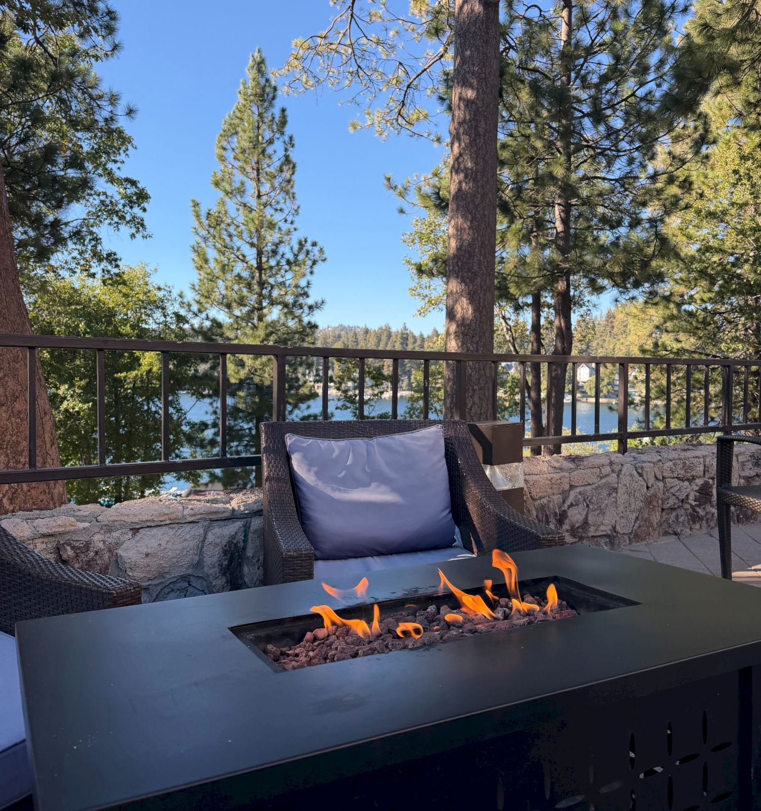 A patio fireplace flickers as tall pine trees frame a lakeside view under a clear blue sky.