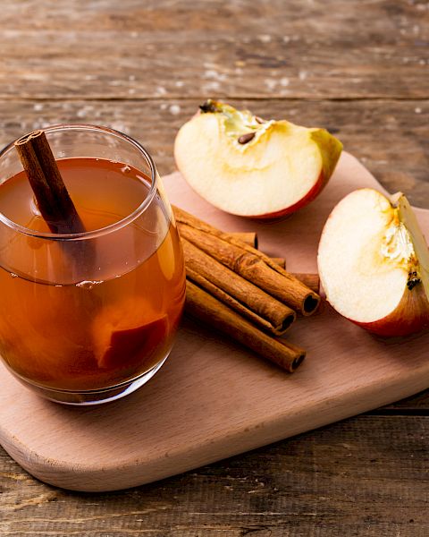 A glass mug of spiced apple cider or tea sits on a wooden board with apple slices and cinnamon sticks.