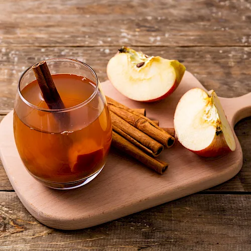 A glass mug of spiced apple cider or tea sits on a wooden board with apple slices and cinnamon sticks.
