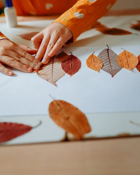 A child&rsquo;s hands arrange colorful autumn leaves on a table, crafting a leaf collage for fall.