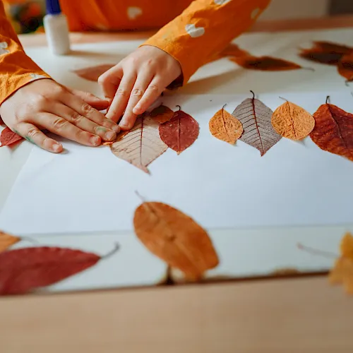 A child’s hands arrange colorful autumn leaves on a table, crafting a leaf collage for fall.