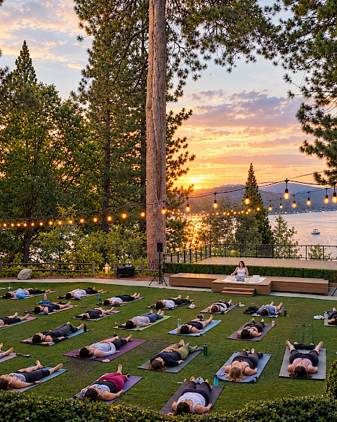 Outdoor yoga on green mats at sunset by a lake, with string lights overhead and people lying in relaxation poses.