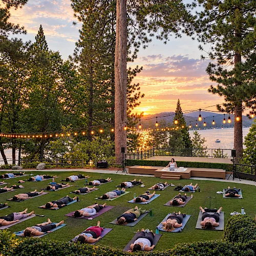 Outdoor yoga on green mats at sunset by a lake, with string lights overhead and people lying in relaxation poses.