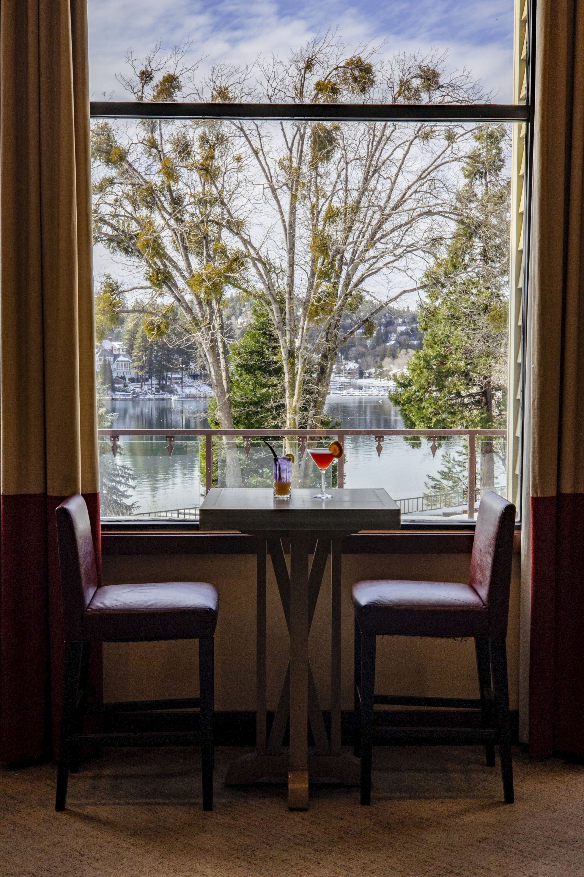Cozy table by a window with cocktails and glasses, looking out over a calm lake lined with trees and greenery outside.