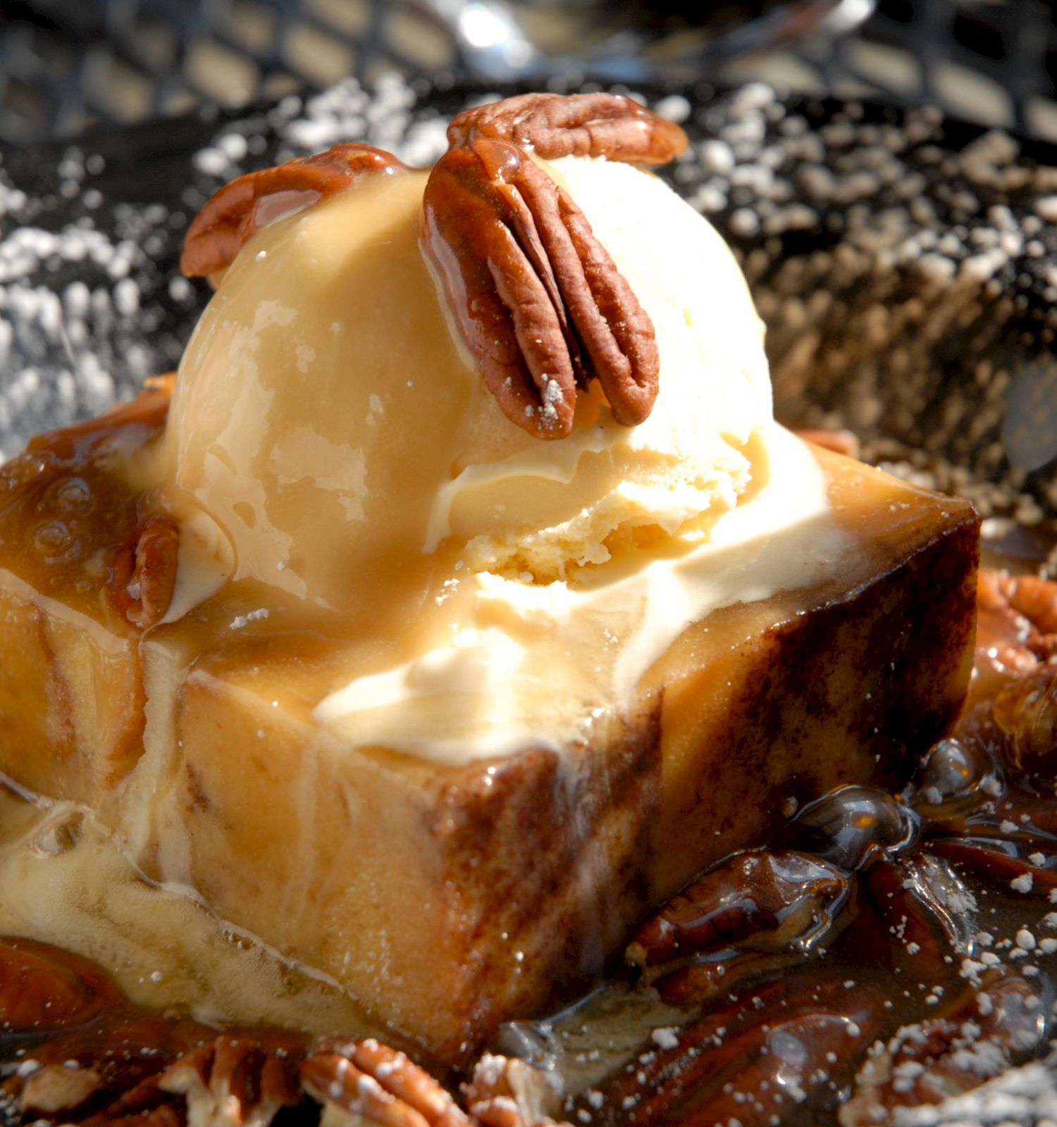 A gooey caramel-topped dessert slice with pecan halves and creamy filling sits on a hot griddle, glistening in the light.