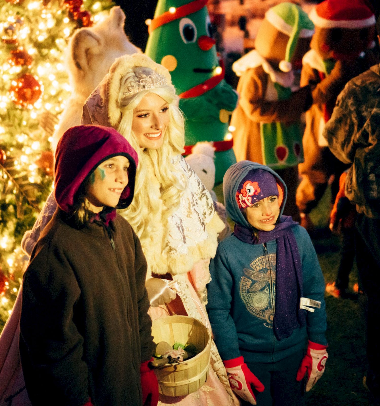 A family in winter clothes and colorful hats stands by a glowing Christmas tree, celebrating with warm holiday cheer.