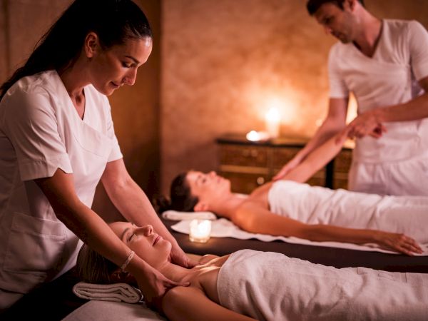 Two people receiving massages in a dimly lit spa, with massage therapists attending to them, enhancing relaxation and well-being.