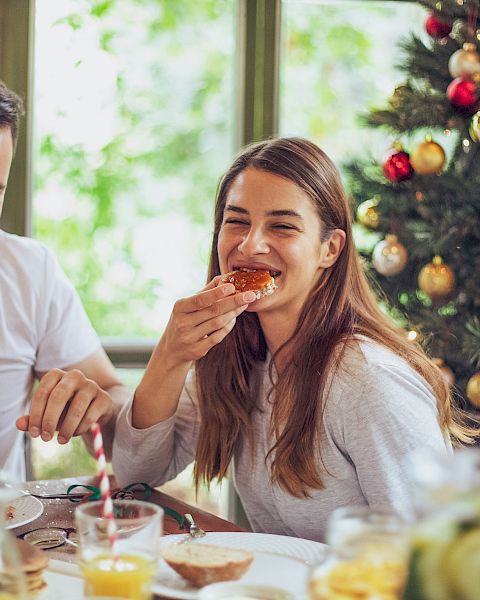 A couple enjoys Christmas breakfast together, smiling as they taste sweet treats by a decorated tree full of red and gold ornaments.