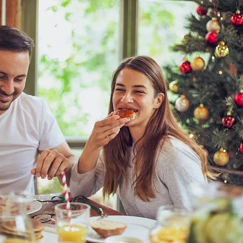 A couple enjoys Christmas breakfast together, smiling as they taste sweet treats by a decorated tree full of red and gold ornaments.