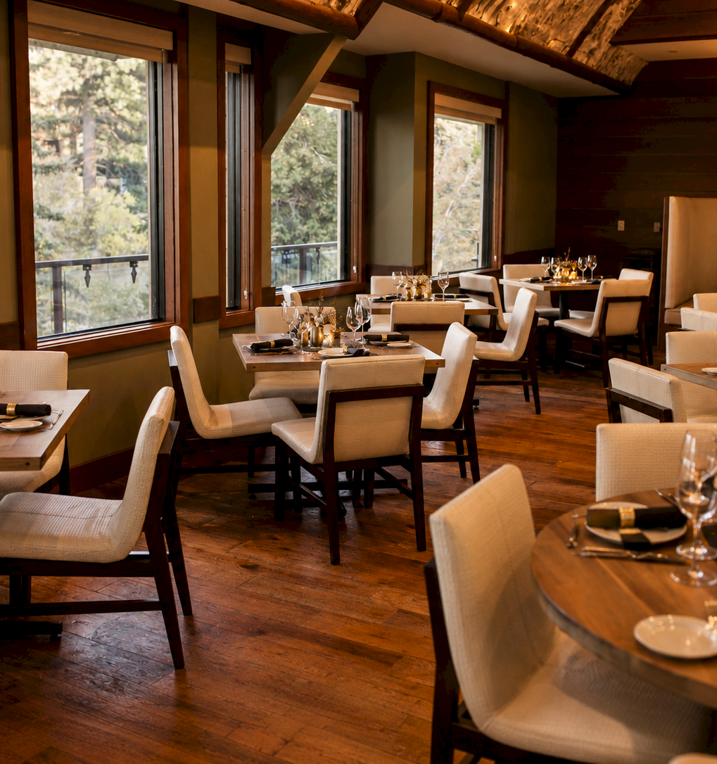 A warmly lit dining room with multiple banquet tables set for a dinner, complete with glassware, candles, and cutlery.