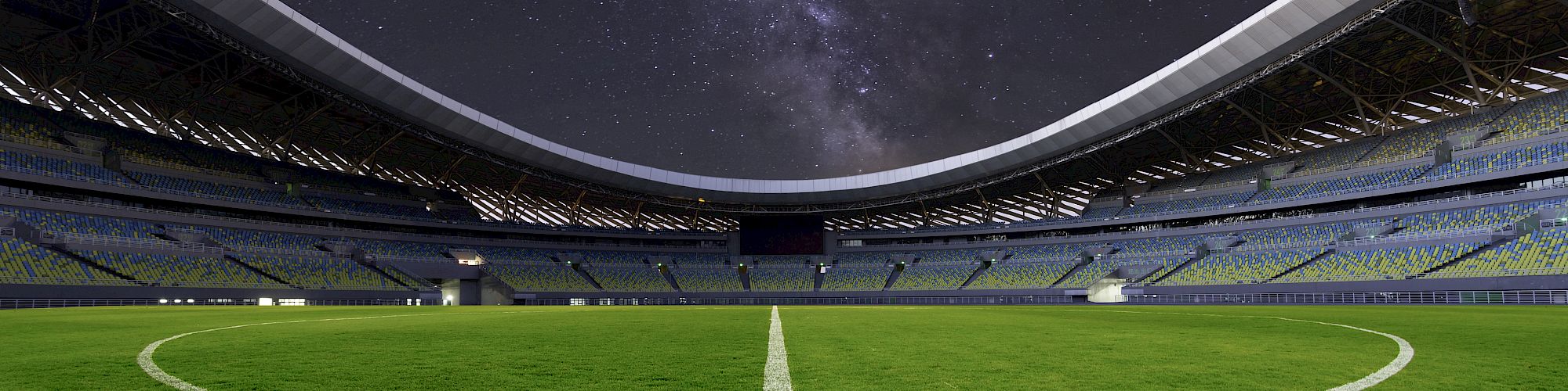 A football stadium at night, empty stands, and a clear starry sky above, with the field illuminated and the center line visible.