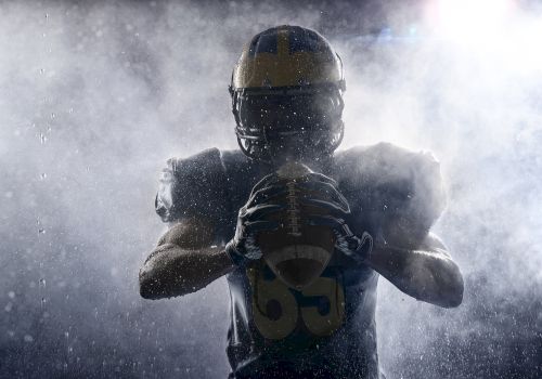 The image shows a football player wearing a helmet, holding a football, with dramatic lighting and mist surrounding them.
