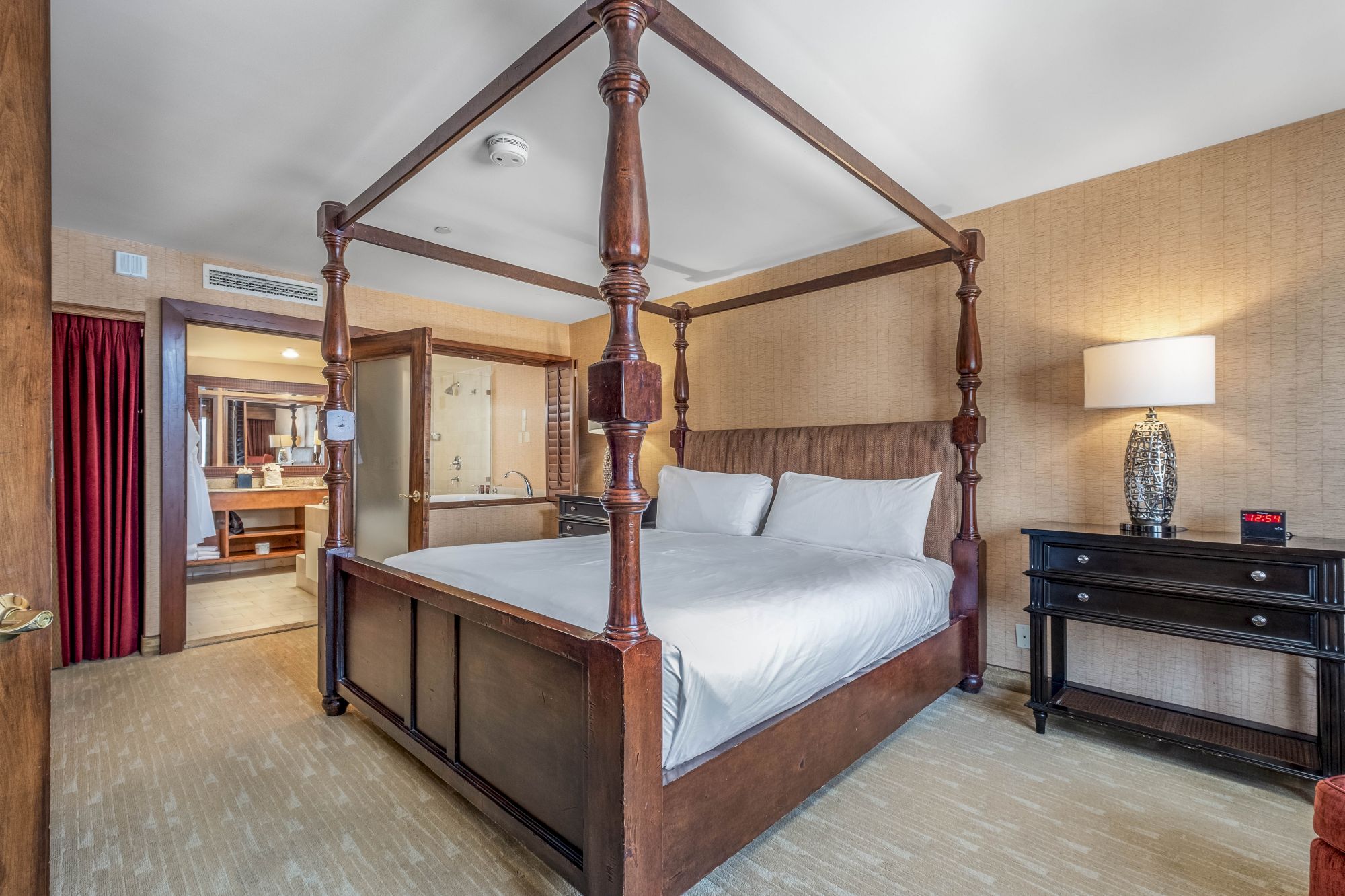 A four-poster bed in a modern hotel room, with wood beams, bedside lamp, and a dresser.