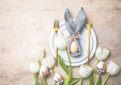 A table setting with a plate, gray napkin shaped like bunny ears, an egg, gold cutlery, decorated eggs, and white tulips is displayed.