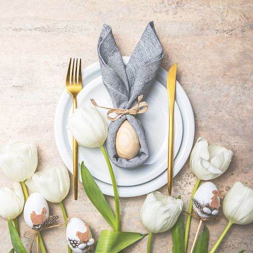 A table setting with a plate, gray napkin shaped like bunny ears, an egg, gold cutlery, decorated eggs, and white tulips is displayed.