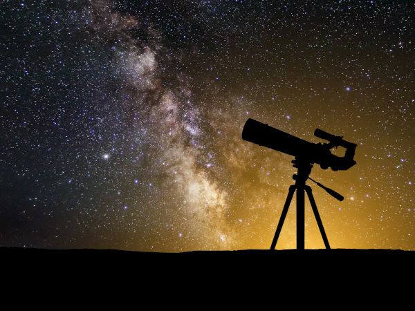 A silhouette of a telescope stands against the starry sky, with the Milky Way galaxy visible in the background.