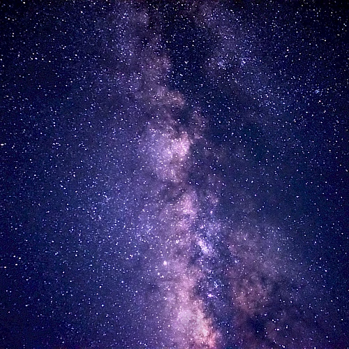 A vibrant night sky with the Milky Way galaxy visible, showcasing numerous stars against a dark, purple-toned backdrop.