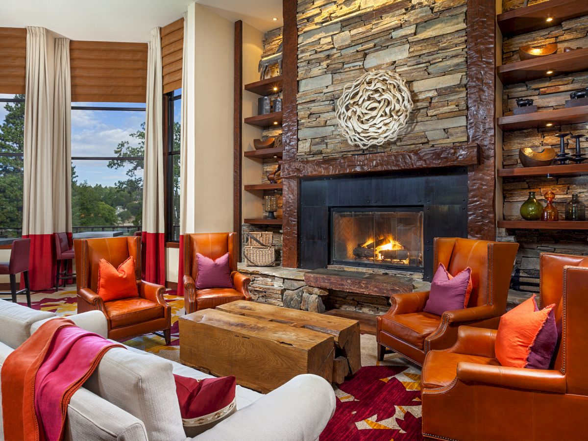 Cozy living room with orange armchairs, stone fireplace, wooden coffee table, colorful accents, and large windows offering a scenic view.