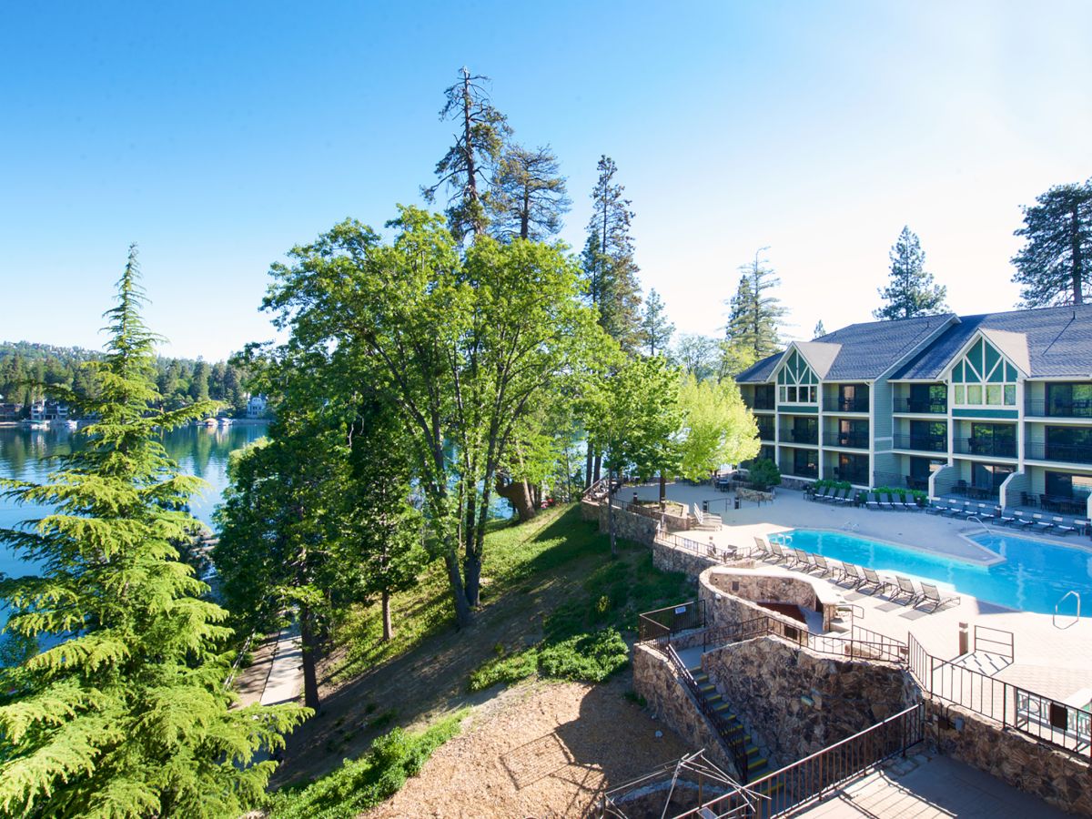The image shows a lakeside resort with a pool, surrounded by trees and buildings, under a clear blue sky.