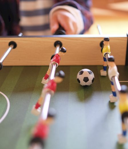 The image shows a foosball table with players in yellow and red uniforms, a ball in the center, and someone playing.
