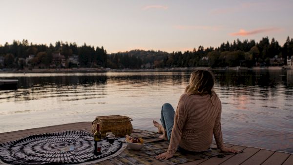 A person sits on a lakeside dock with a picnic setup, enjoying a tranquil sunset view over the water, surrounded by trees.