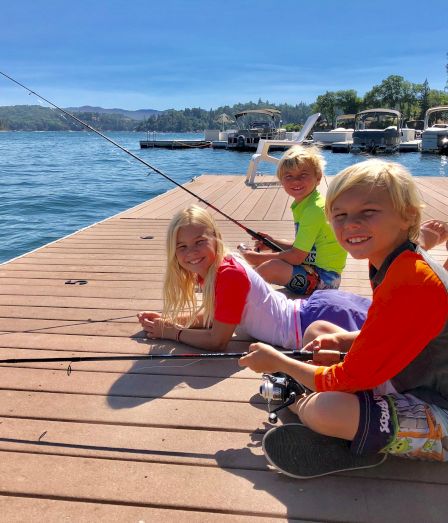 Three kids smile on a lake dock with a fishing rod, with boats and a sunny shoreline in the background.