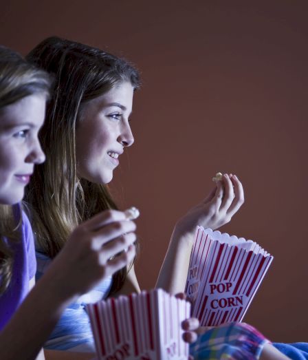 Two people are watching something, holding popcorn boxes, in a dimly lit room.