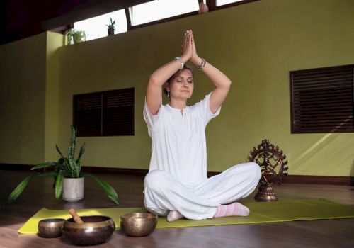 A person in white practices meditation on a yoga mat indoors, with a plant, singing bowls, and a statue nearby.