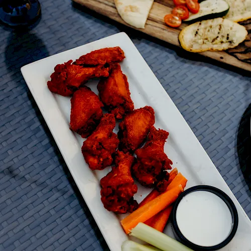 A platter of spicy red fried chicken with carrot/celery sticks and a side of white dip, plus fries and flatbread in the background.